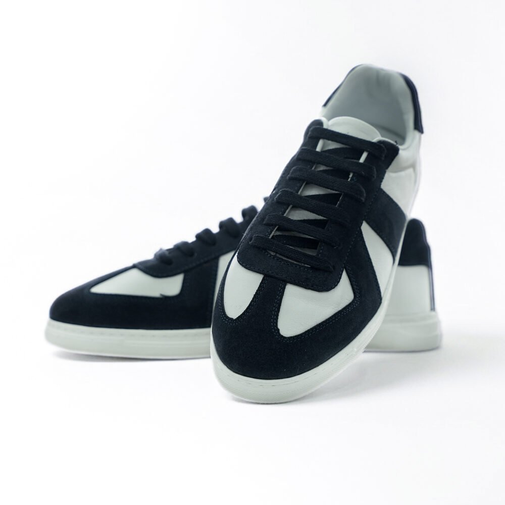 Beyaz deri ve lacivert süet panelli low-top sneaker çiftinin önden ve açılı şekilde konumlandırıldığı dinamik ürün çekimi